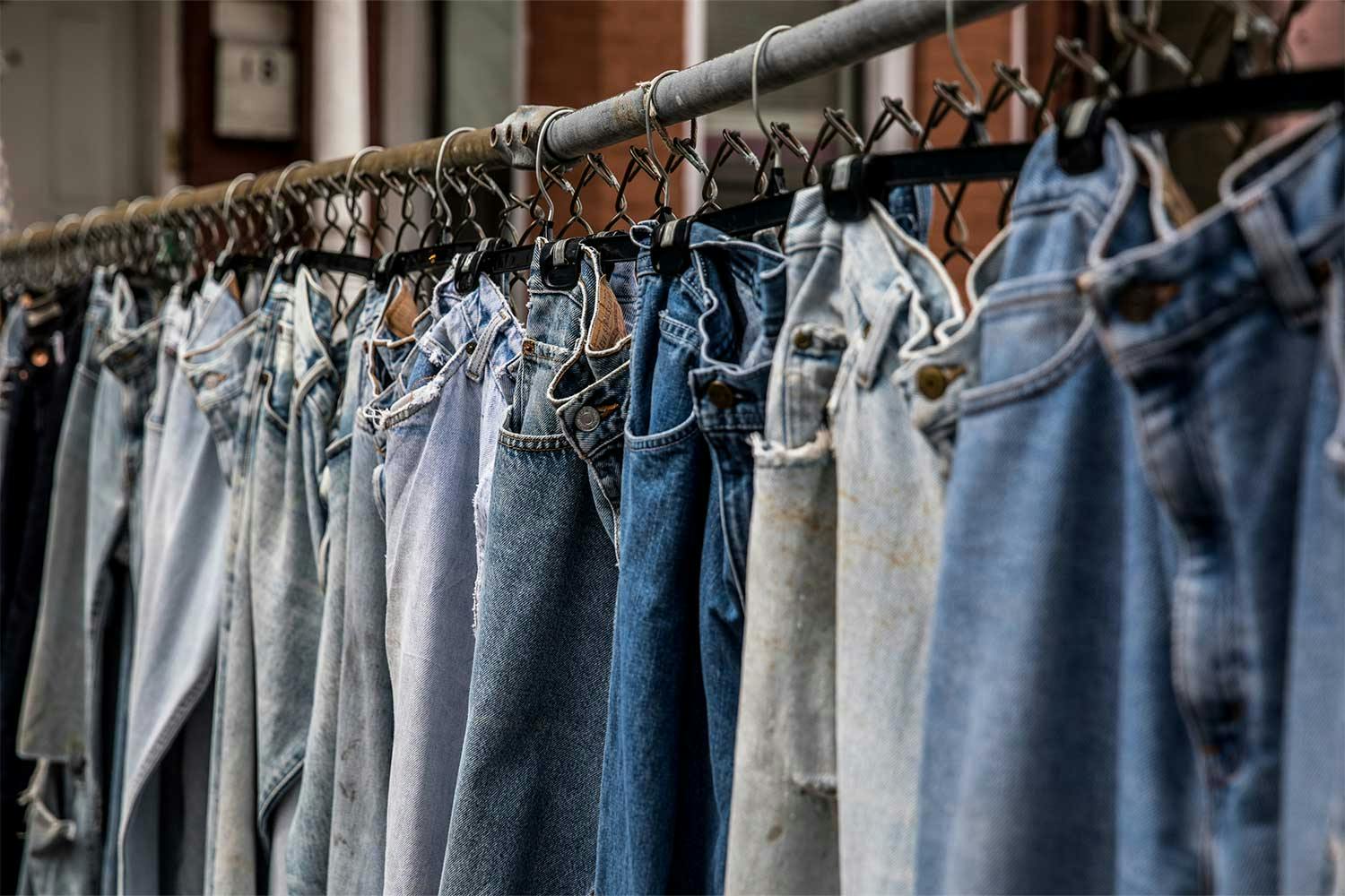 Jeans in various colours hang on a chain link fence in front of a red brick building.