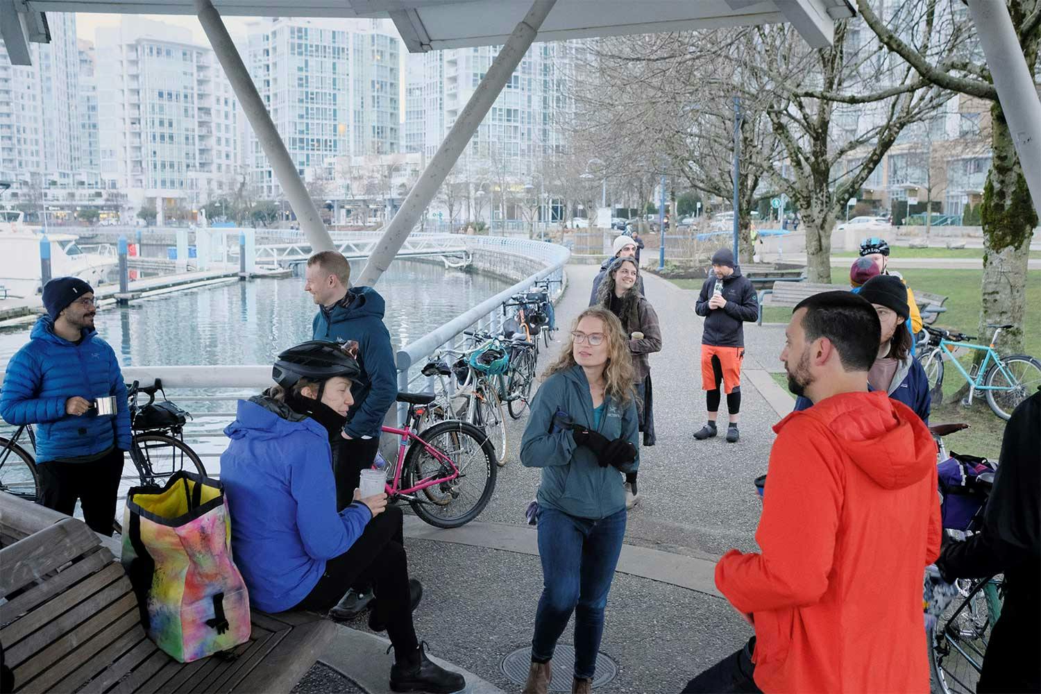 A group of cyclists drink coffee at a harbourfront park in Vancouver, their bikes parked against railings and trees nearby.