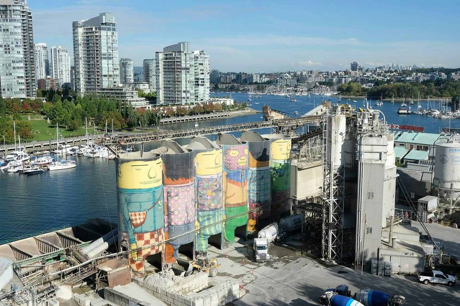 Murals of giant people on 6 silos at a waterfront concrete facility. In the background are a park and apartment towers.