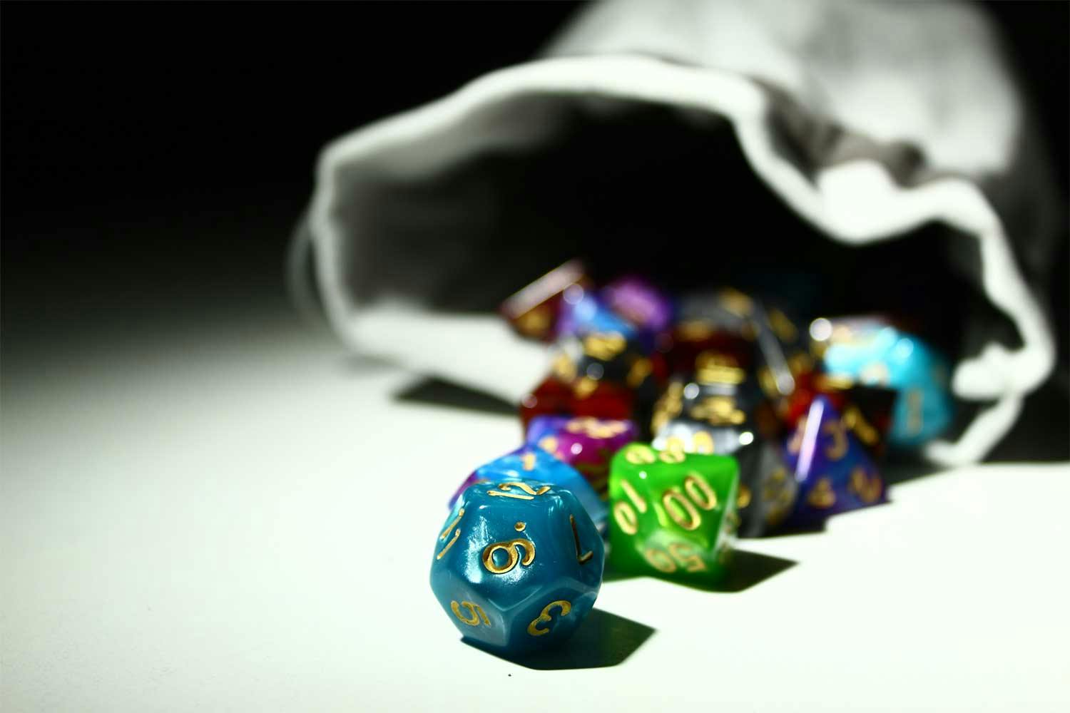 Several multi-sided game dice spill out of a grey cloth bag onto a white tabletop.