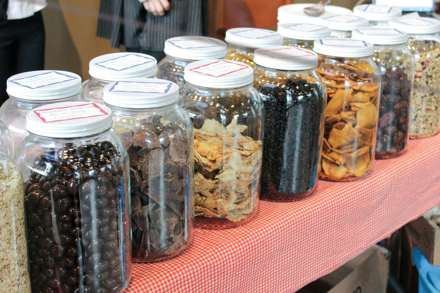 Two rows of glass jars containing dried fruit, nuts, chocolate, and grains on a red checkered tablecloth.