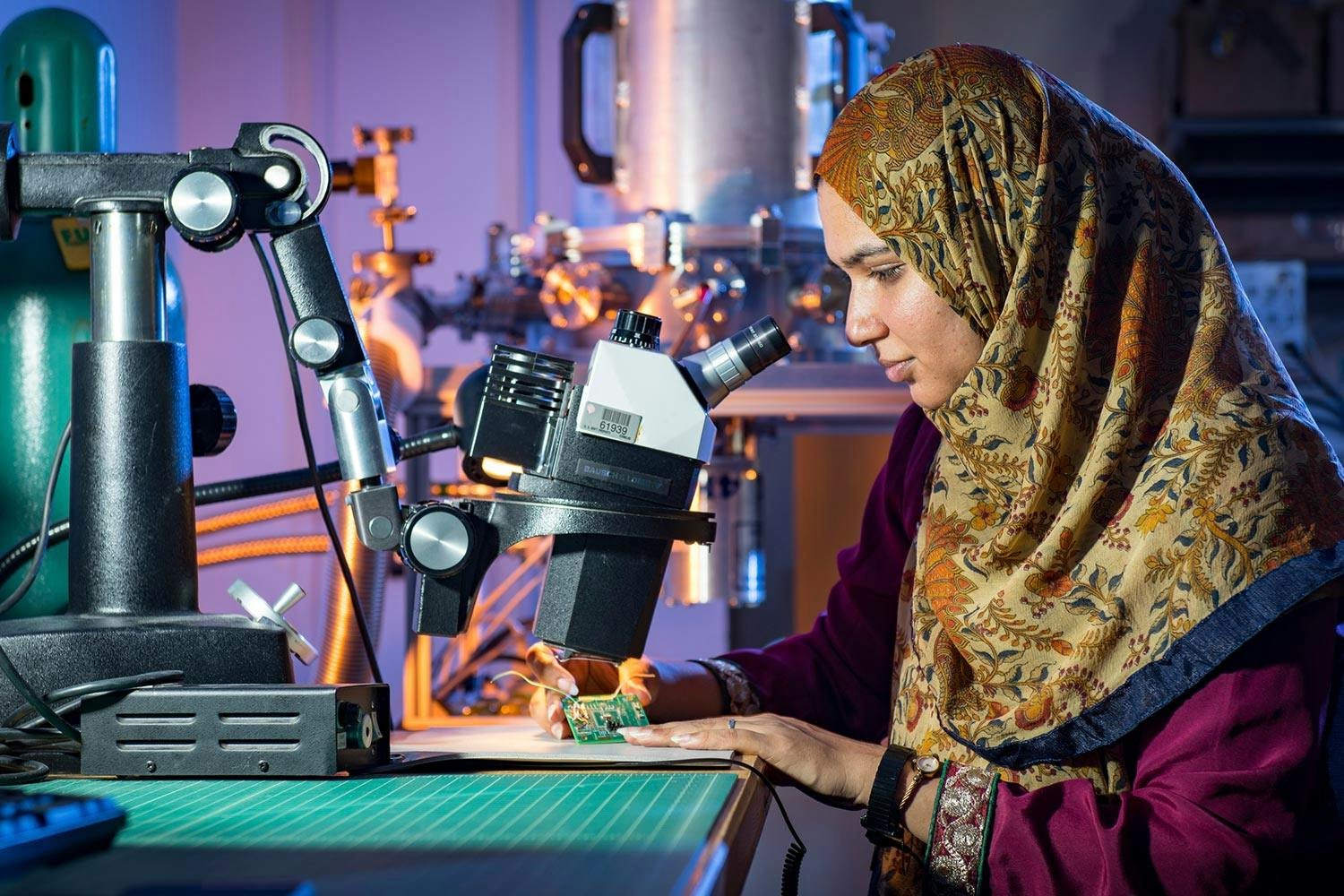 A woman with brown skin wearing a khaki floral hijab holds a circuit board under a microscope, looking into the eyepiece.