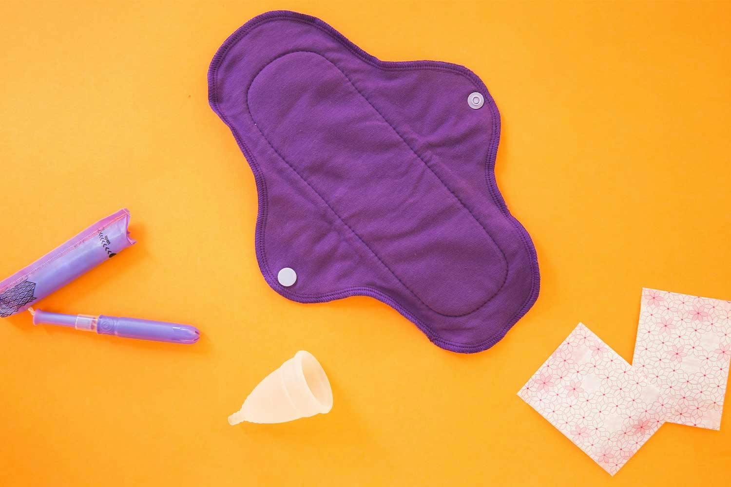 A purple fabric reusable menstrual pad, tampon, clear plastic menstrual cup, and disposable pads on an orange background.