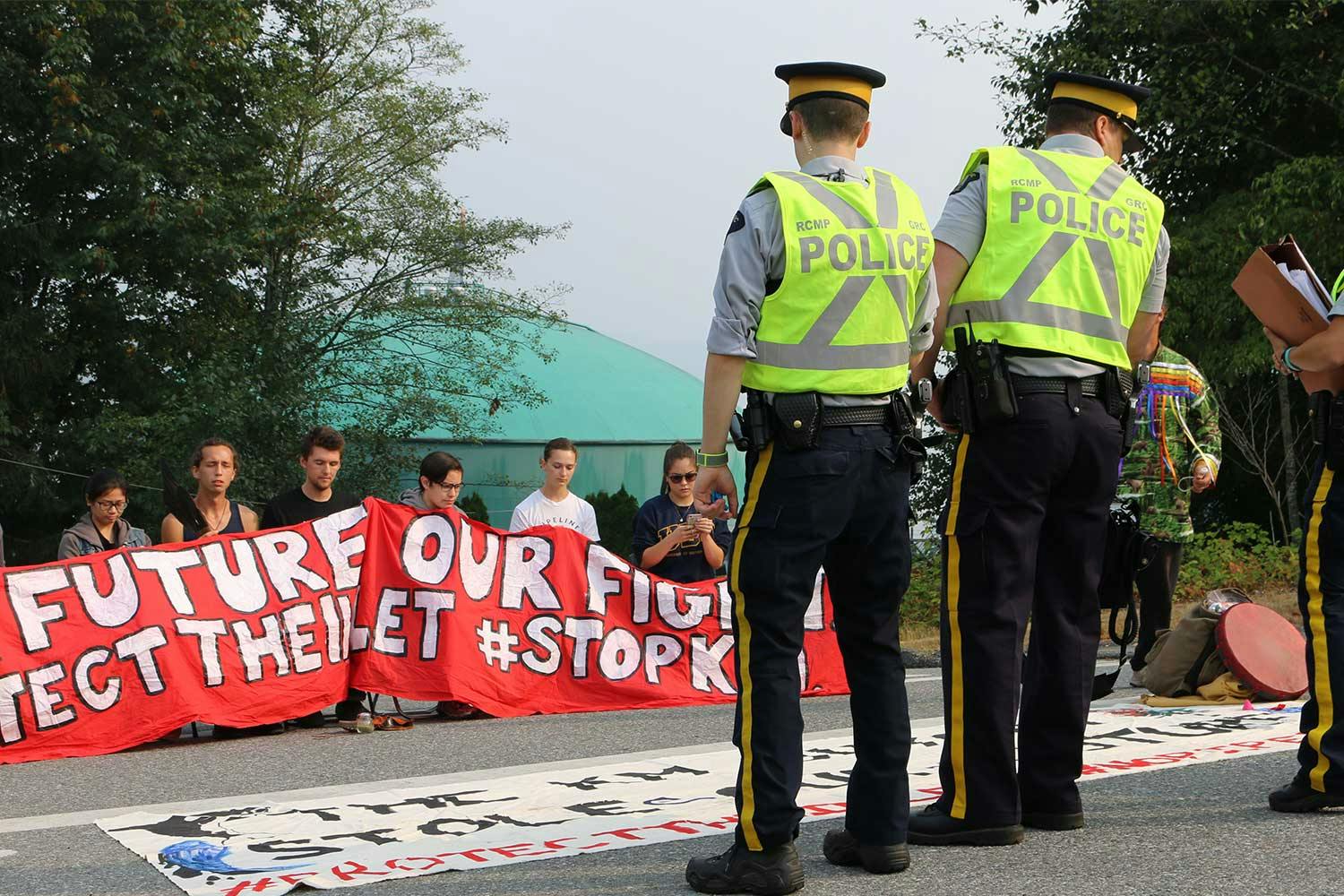 Two police officers in bright yellow vests stand before a group of activists with a red banner that says "Protect the Inlet".