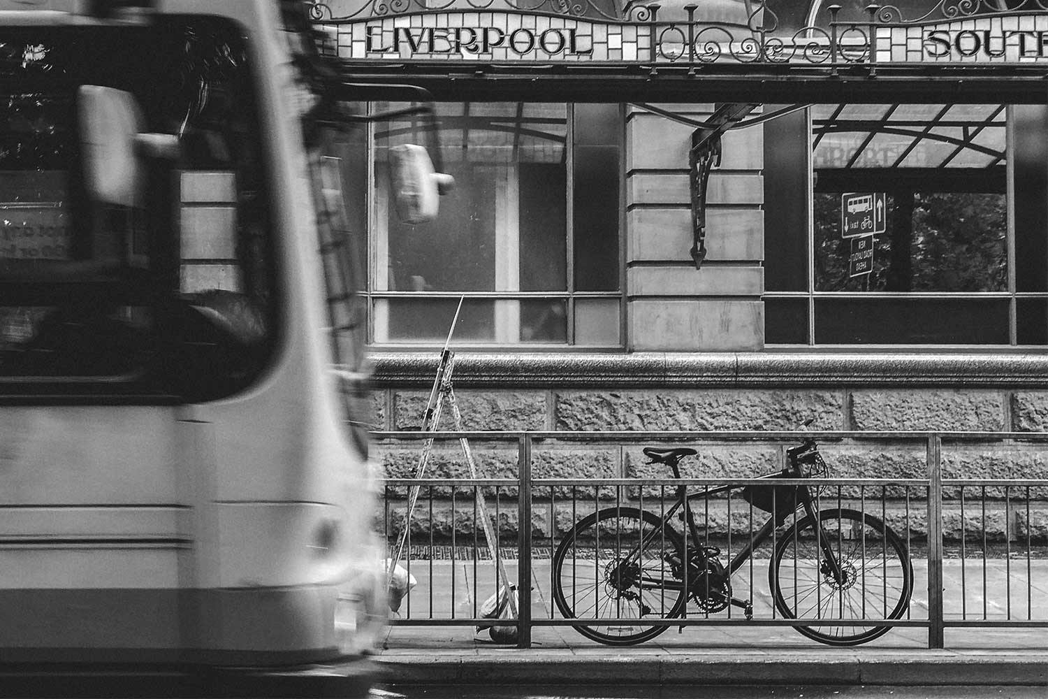 A bike leans against a metal fence as a bus drives past. Behind is a stone building with an awning that reads "Liverpool."