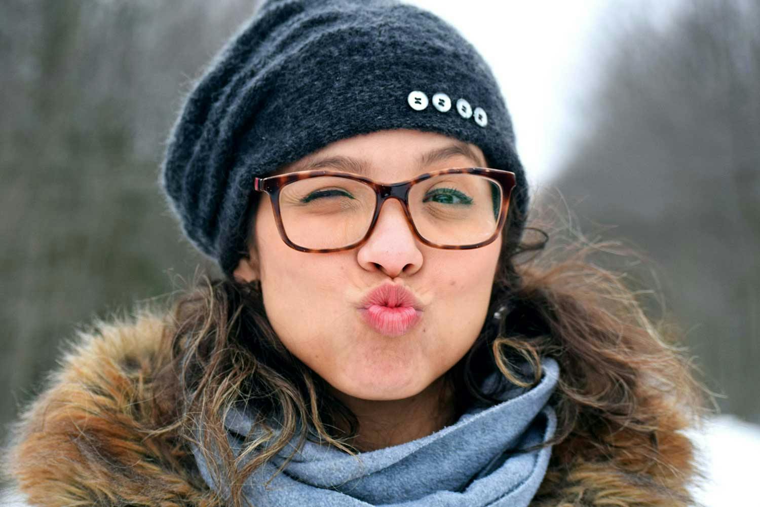A light-skinned woman wearing glasses, a grey knit hat, and a fur-lined jacket winks and purses her lips.