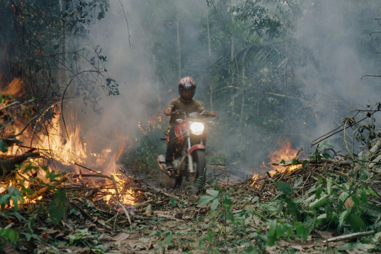 A helmeted man rides a red motorbike through a smoky, burning patch of the Amazon Rainforest.