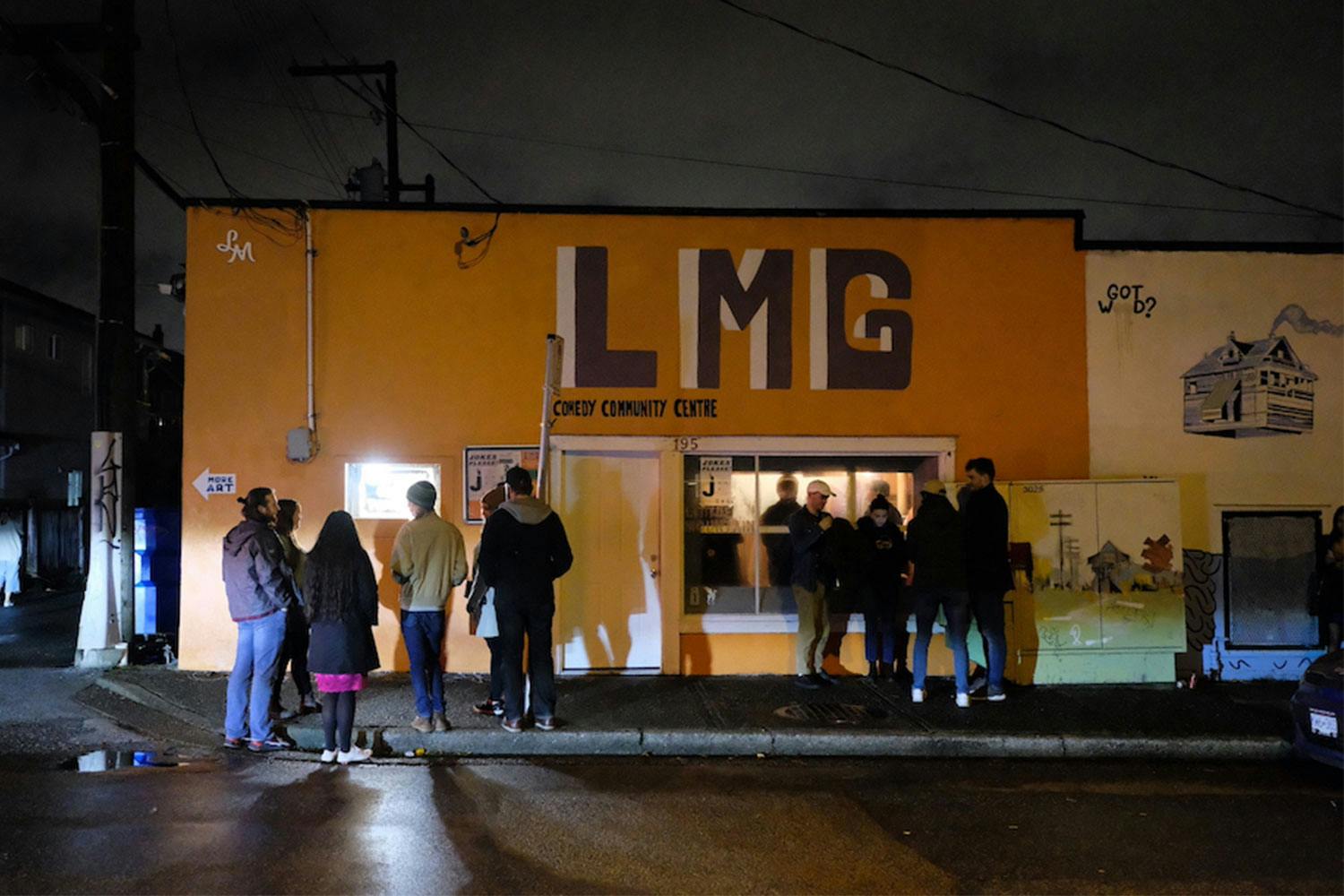 Groups of 4-5 people stand outside an orange building with the letters "LMG" painted in purple above the front door.