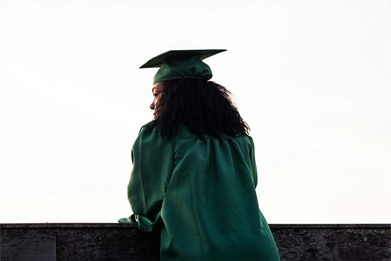 A woman with brown skin, dark hair, wearing a green graduation cap and gown leans against a concrete ledge, looking stoic.