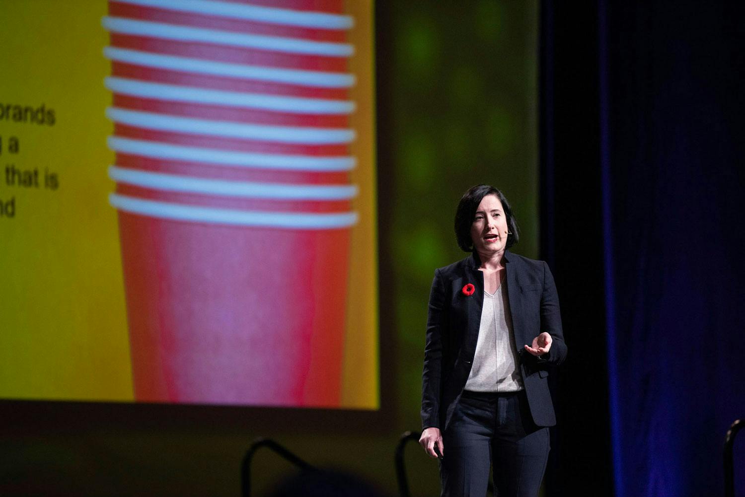Kate Daly, a white-skinned woman with short black hair speaks on stage. On screen behind her is a stack of red plastic cups.