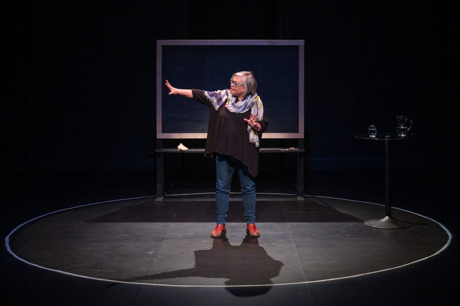 Alanna Mitchell on stage, right hand held out, in front of a chalkboard. She has light skin, short grey hair, and glasses.