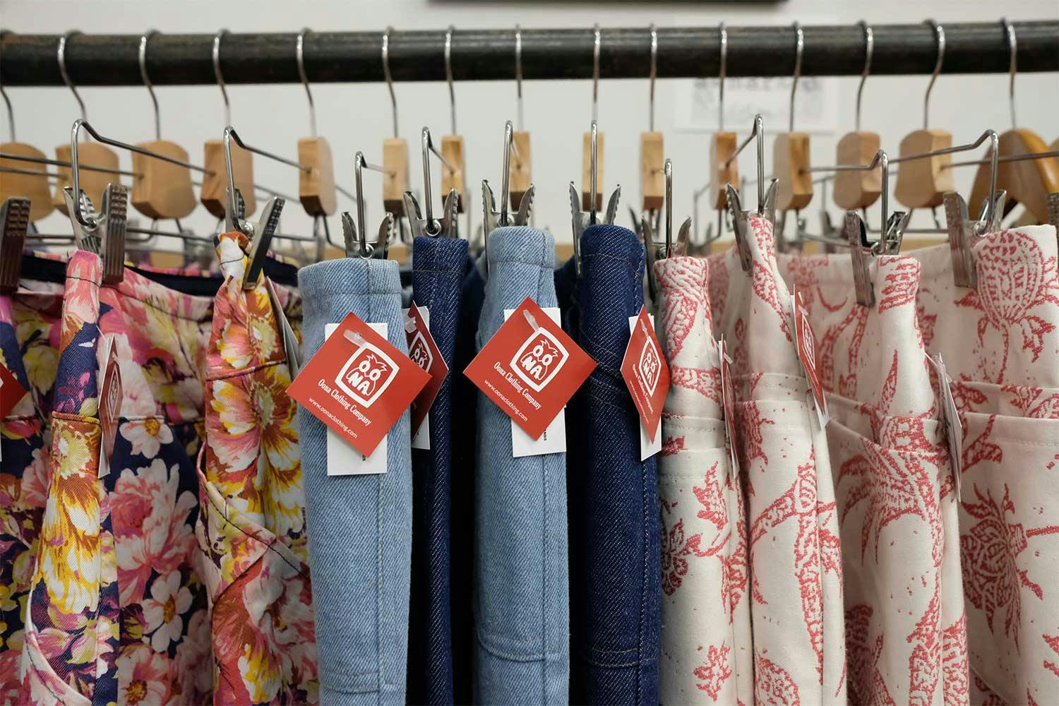 A row floral print and denim pants hanging from hangers. A red label with the Oona Clothing logo appears on each pair.