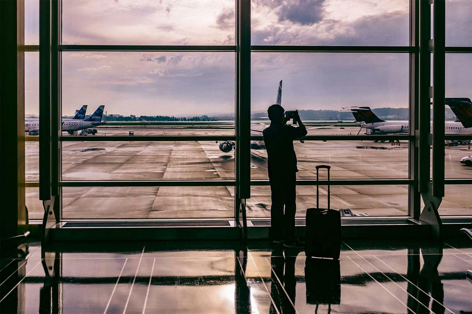 A person uses their phone to take a photo of airplanes sitting on tarmac through a large airport window.