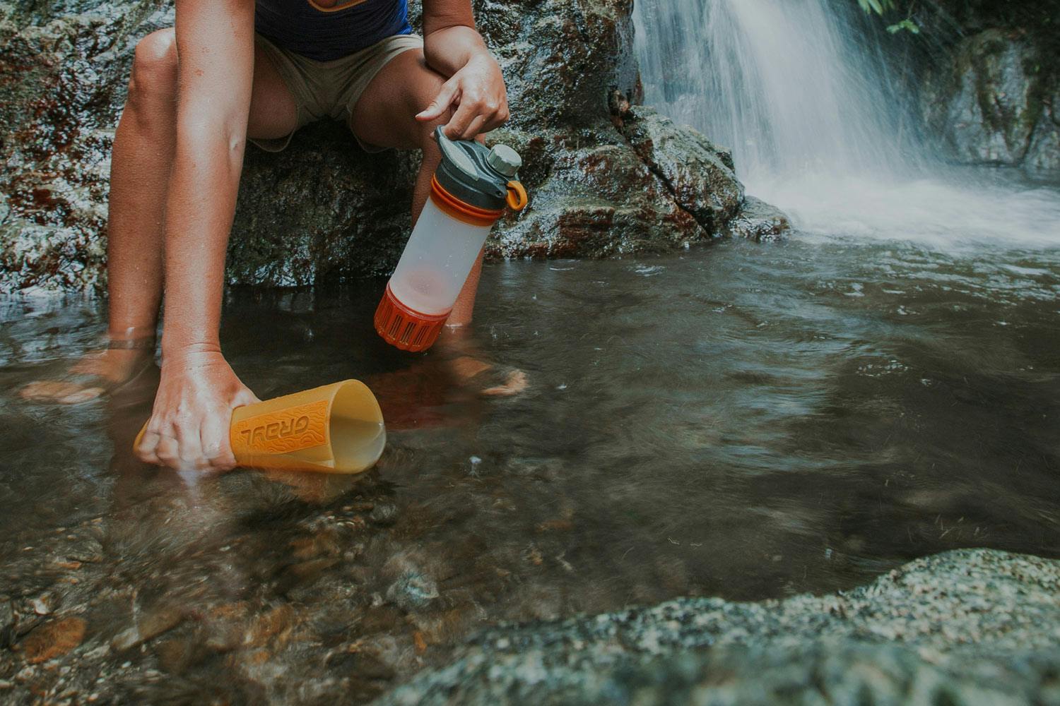 A person sits near a waterfall scooping water into a portable water filtering bottle.