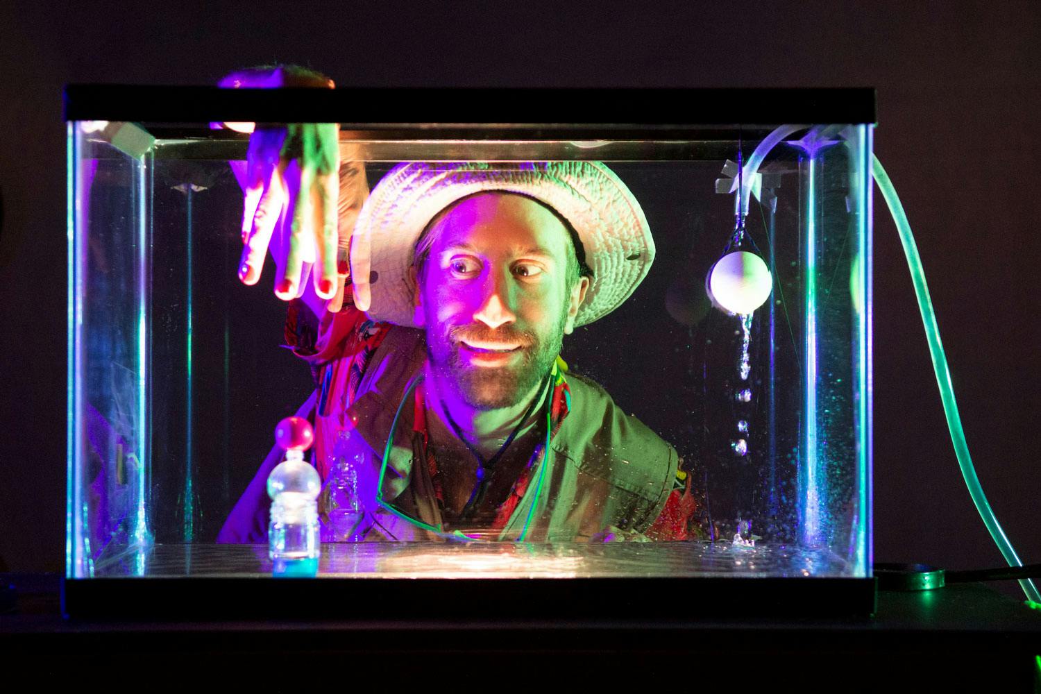 A white man wearing a wide-brimmed hat and green vest reaches his fingers into a glass box. He is lit with pink and green light.