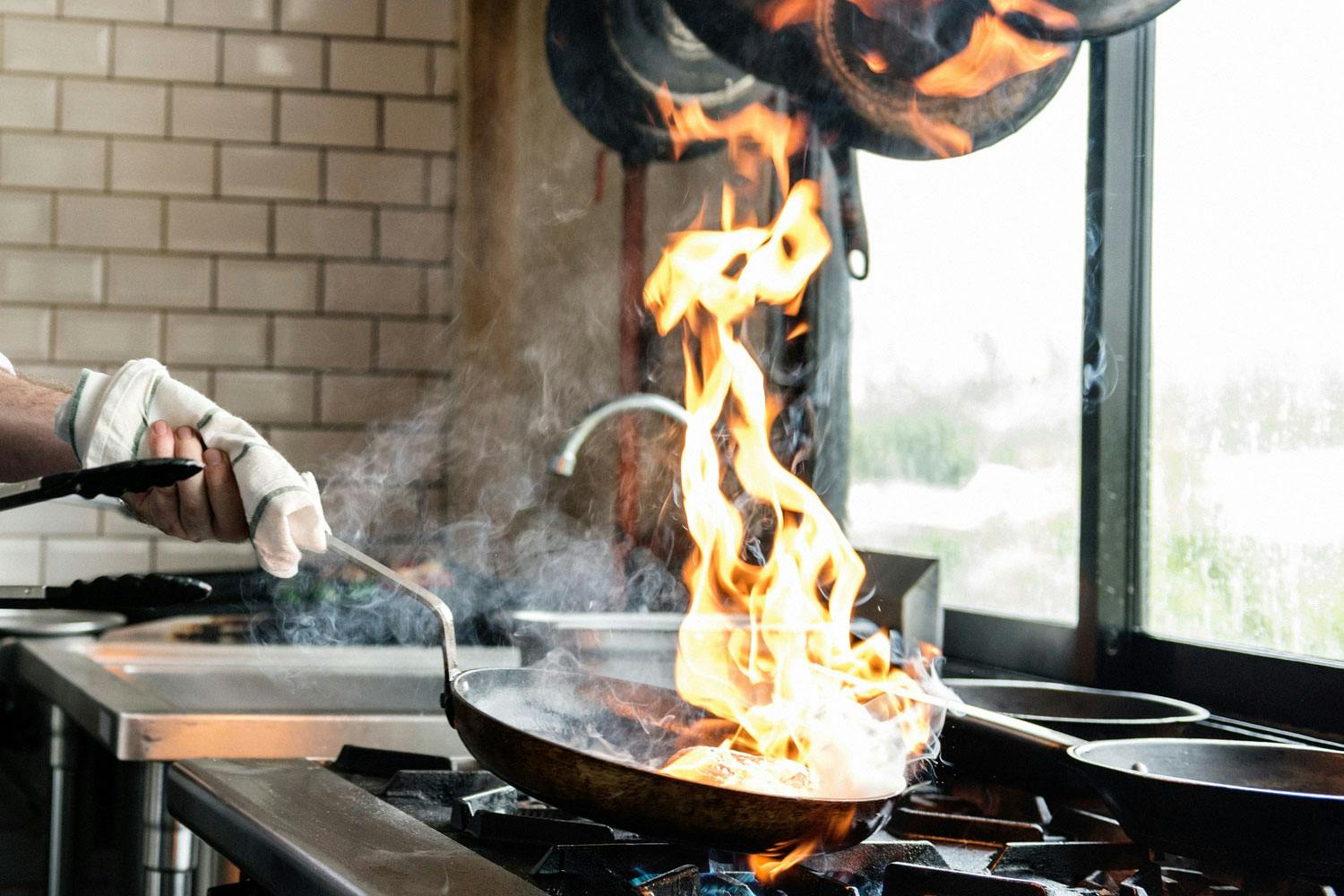 A huge flame rises from a frying pan held above a gas stove inside a restaurant kitchen.