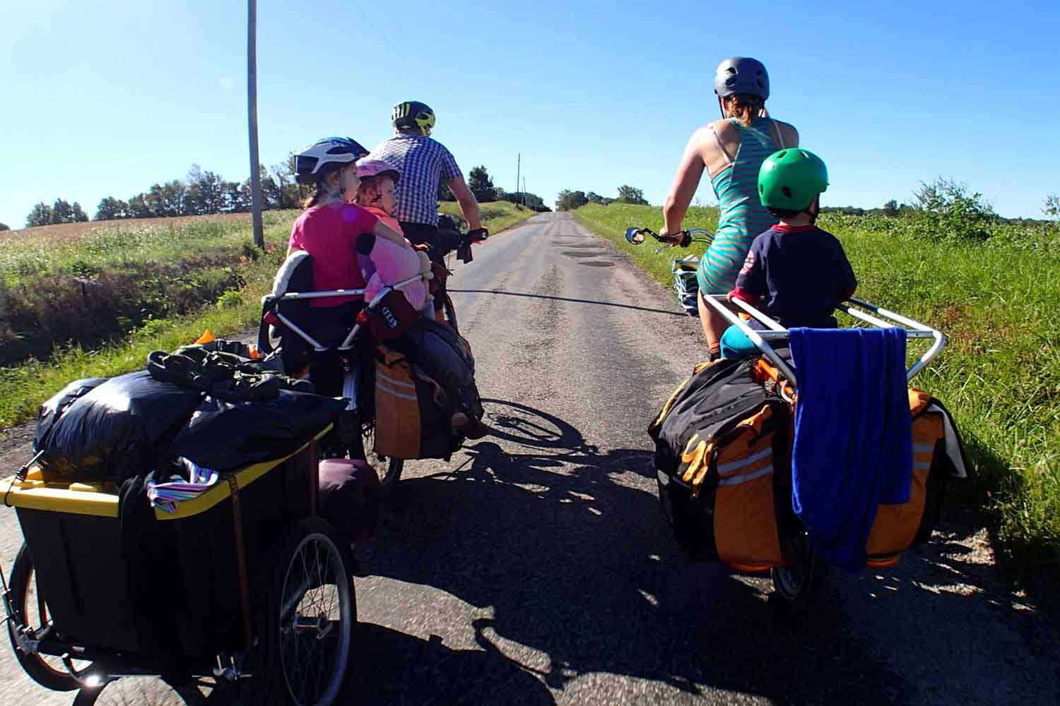 2 parents ride bikes hitched to child seats and cargo. Two kids ride with one parent, and a third kid rides with the other.