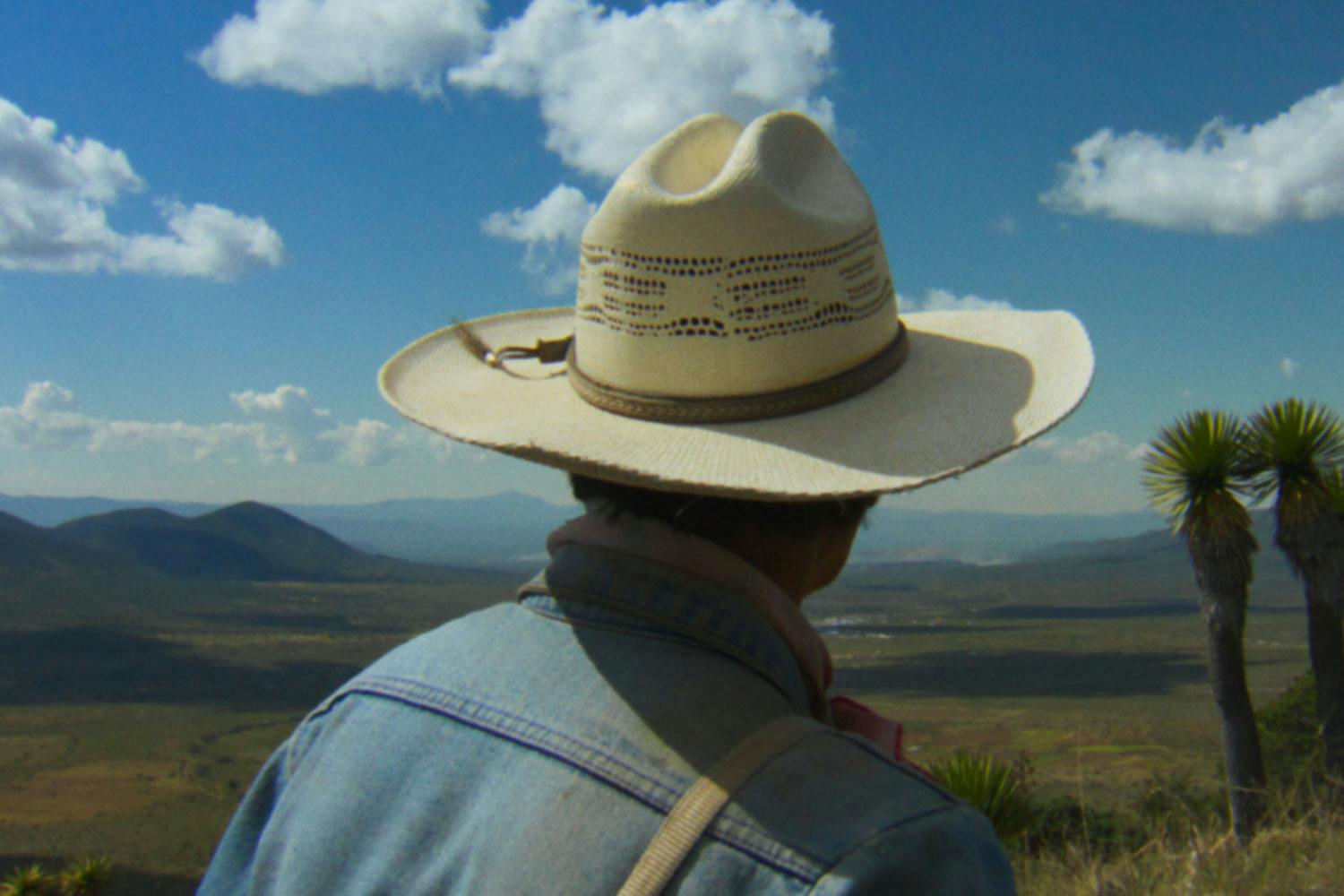Roberto de la Rosa faces away, looking down at a vast, green valley, wearing a wide-brimmed hat and jean jacket.