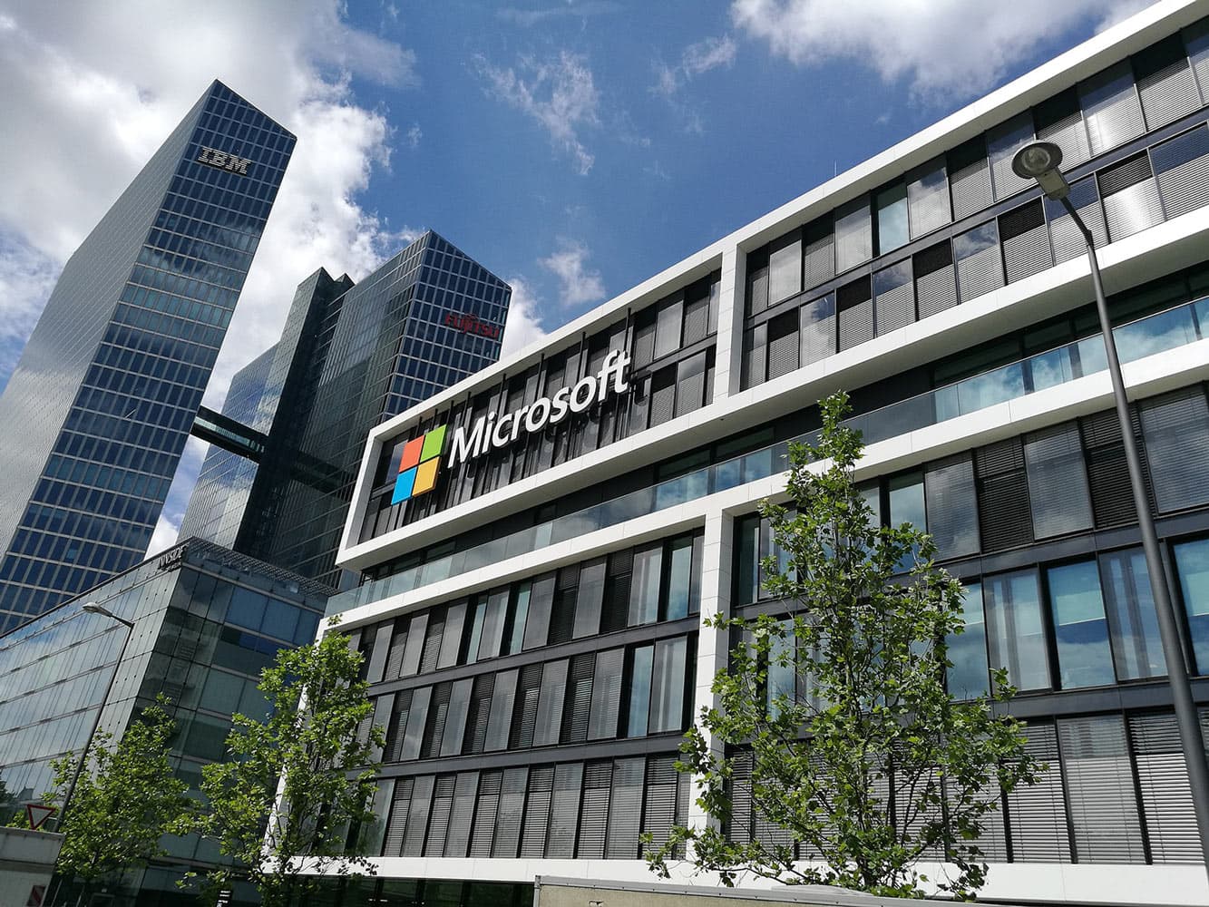 A rectangular glass-covered building with Microsoft's logo in Germany. Behind are 2 towers with the IBM and Fujitsu logos.