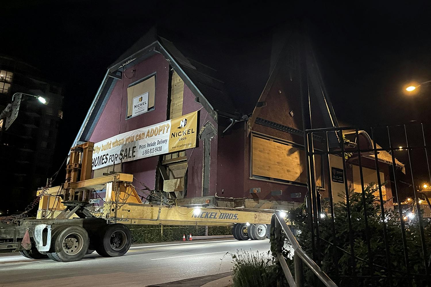 At night, a large burgundy wood-paneled house is loaded onto a large yellow trailer for relocation.