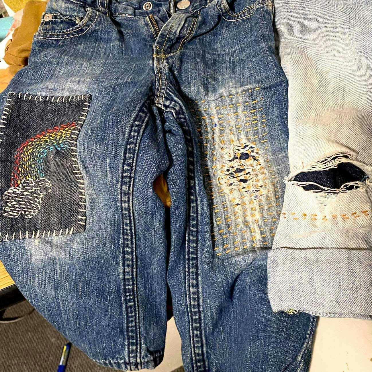 Jeans with mended thighs. One leg has a dark rectangle patch with a stitched rainbow; the other has rows of yellow stitches.