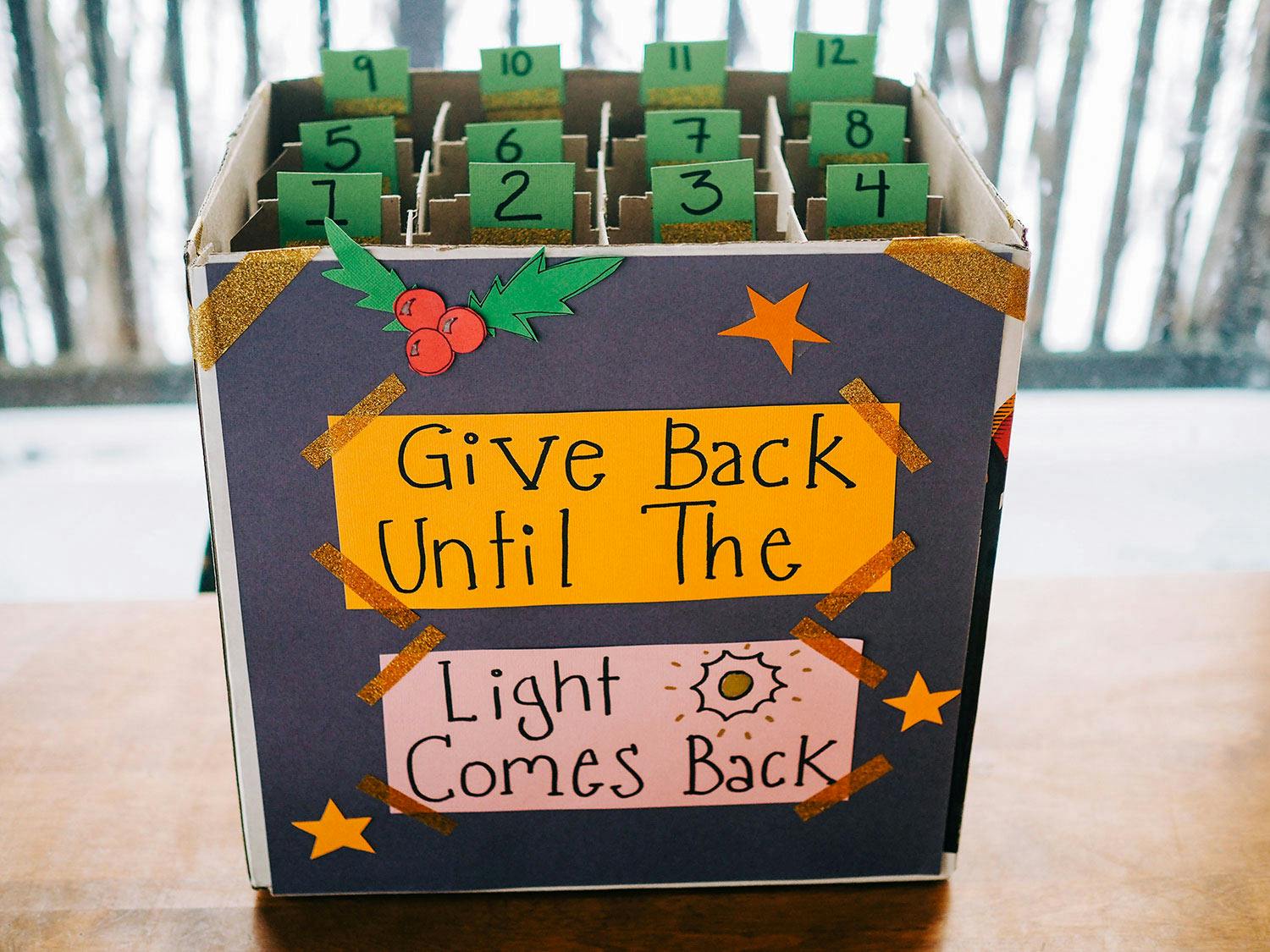A box with 12 advent calendar-like sections inside decorated with paper stars and holly and labelled “Give Back Until the Light Comes Back.”
