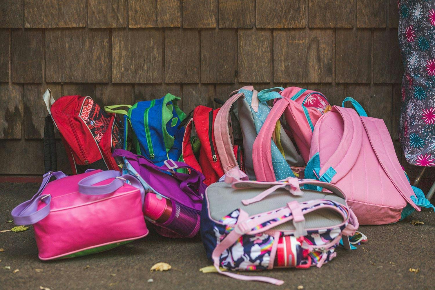 A small pile of colourful children's backpacks sit against a dark brown wood-panelled exterior wall.