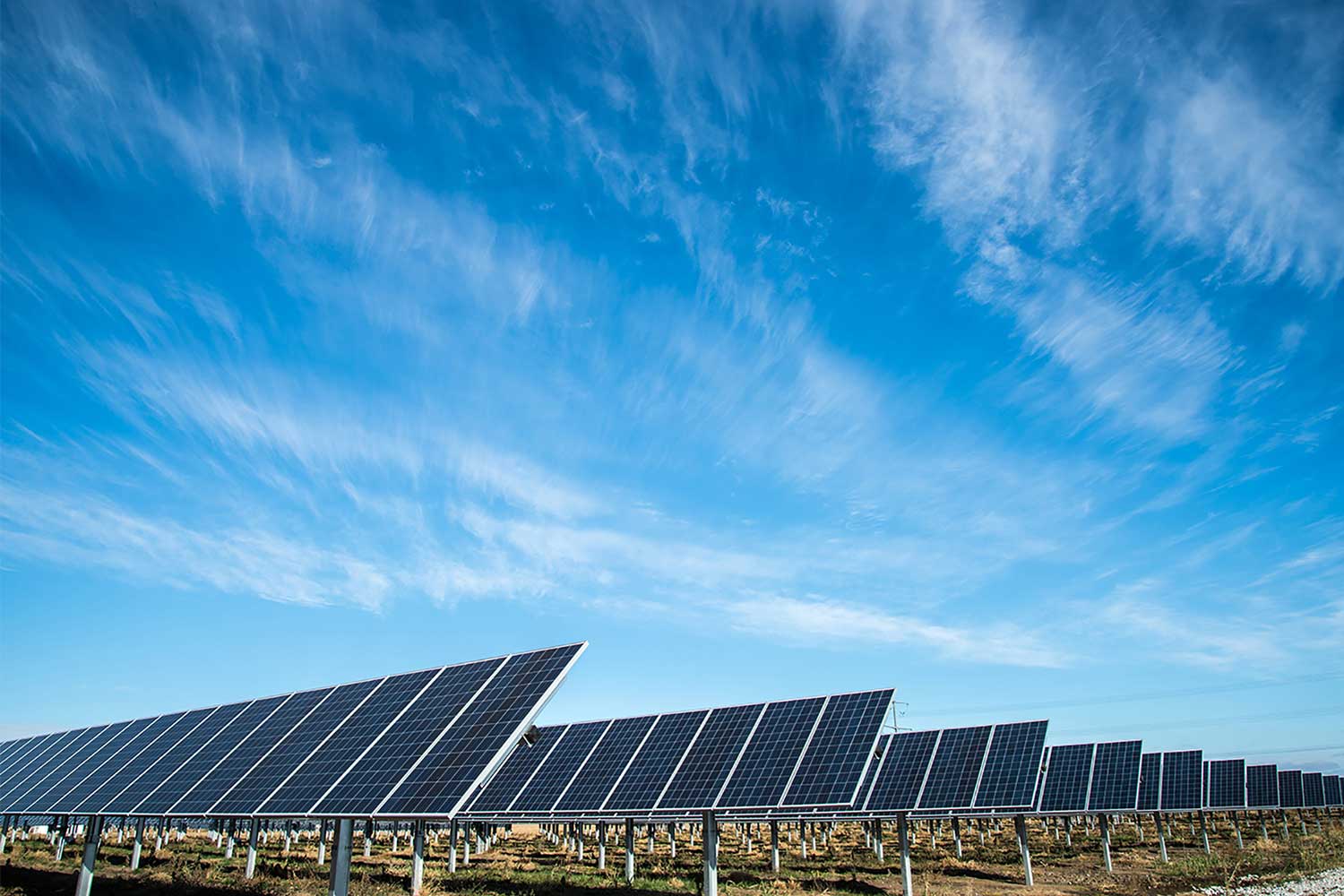 A large array of solar panels on a flat field under a bright blue sky with wispy clouds.