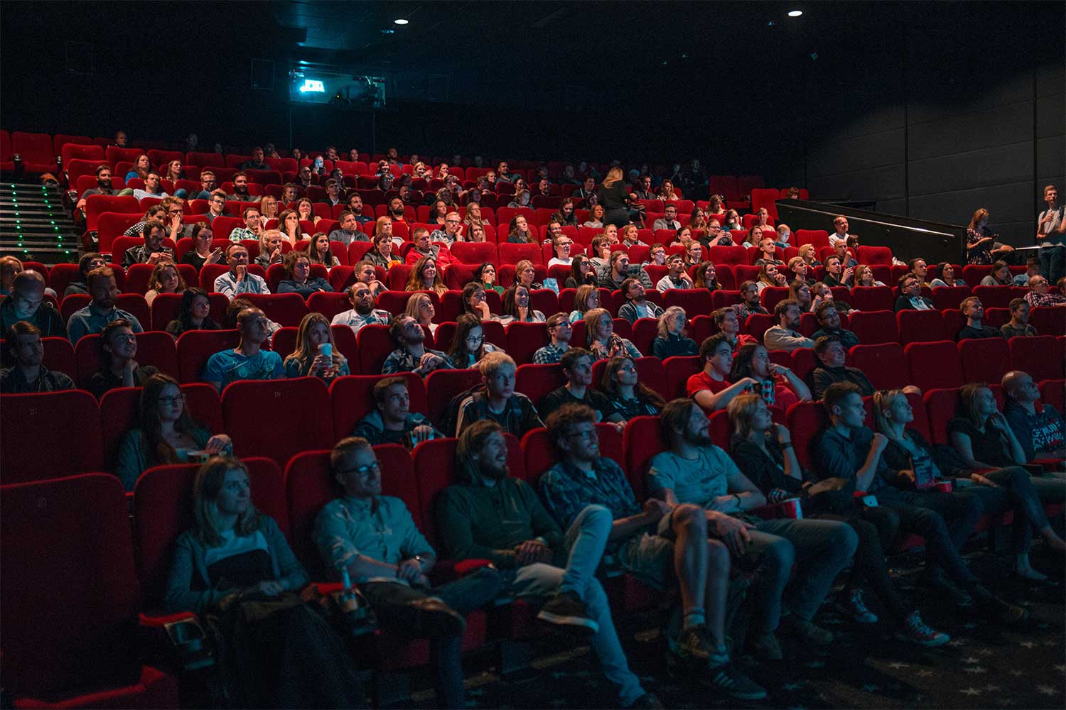 A movie theatre with red seats, seating an almost full audience. The front row is lit softly by the blue light of the screen.