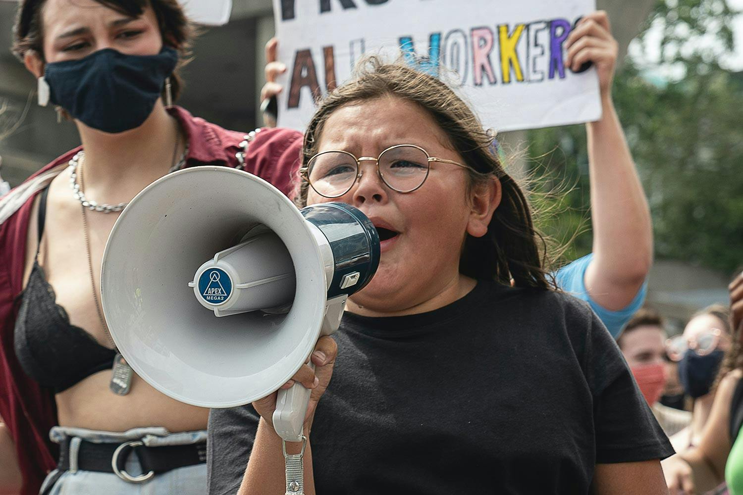 A child with long brown hair, wheatish skin, and glasses shouts into a megaphone as a crowd of protesters walks behind her.