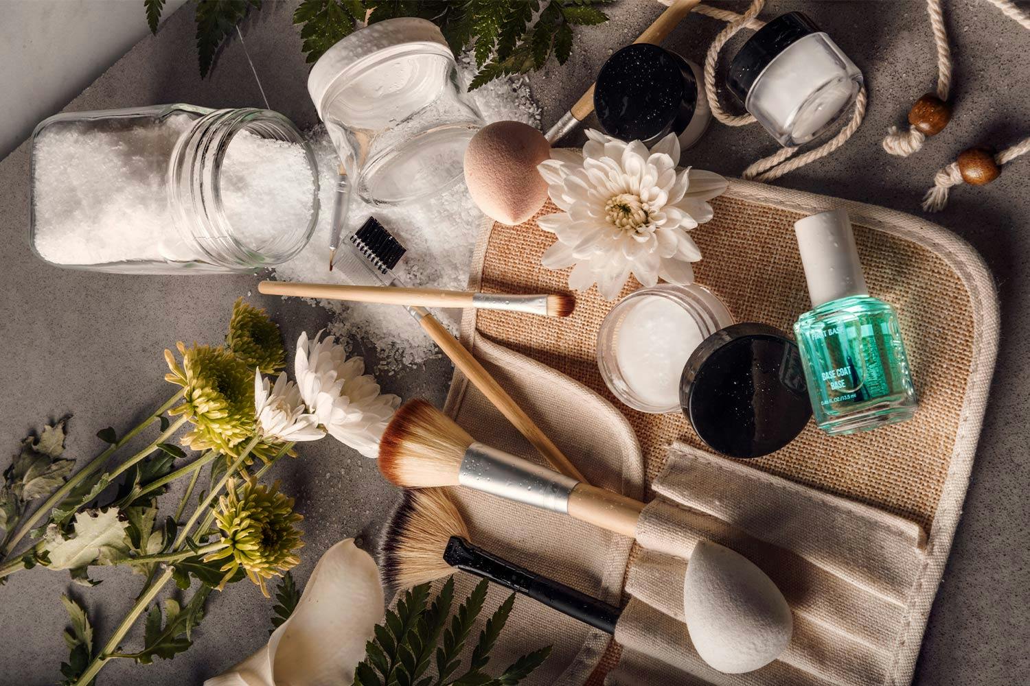 Makeup brushes in a roll-up case, small containers, nail polish, and flowers spread out on a grey table.