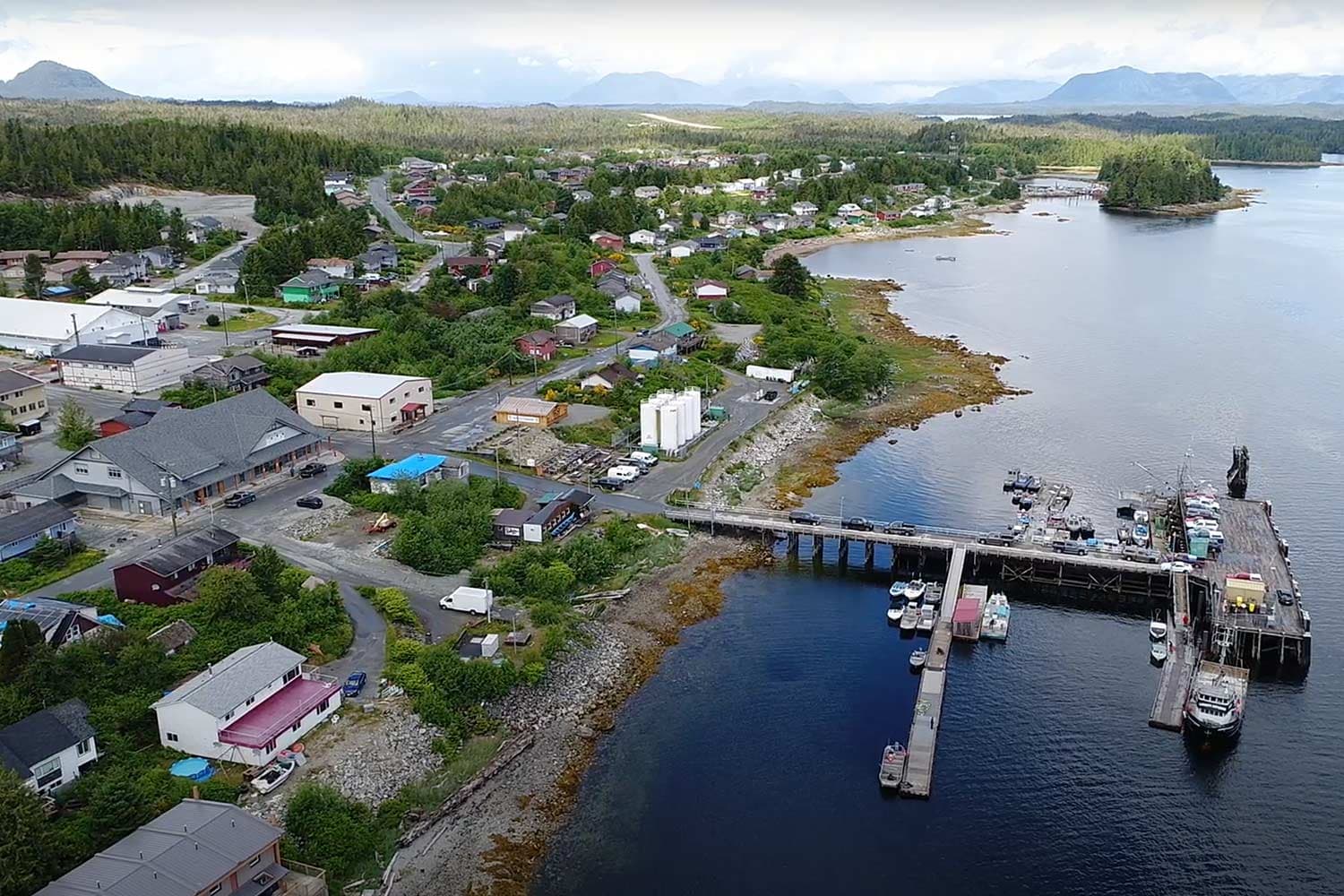Aerial view of Bella Bella, a coastal town. Commercial buildings and homes dot the vast forest and a pier juts off the shore.