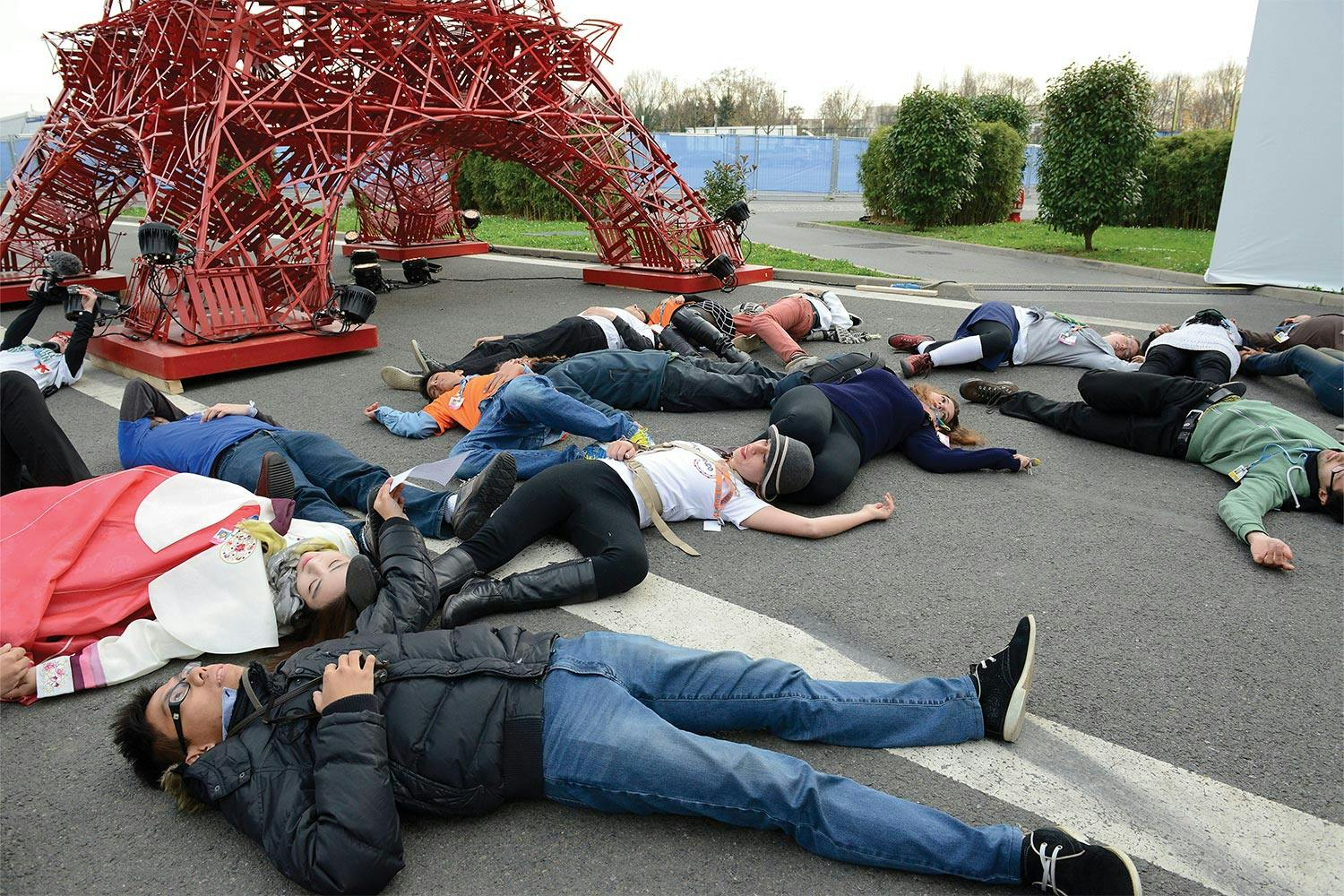 A group of youth lie on the pavement next to a large sculpture of the Eiffel Tower made of red folding chairs.