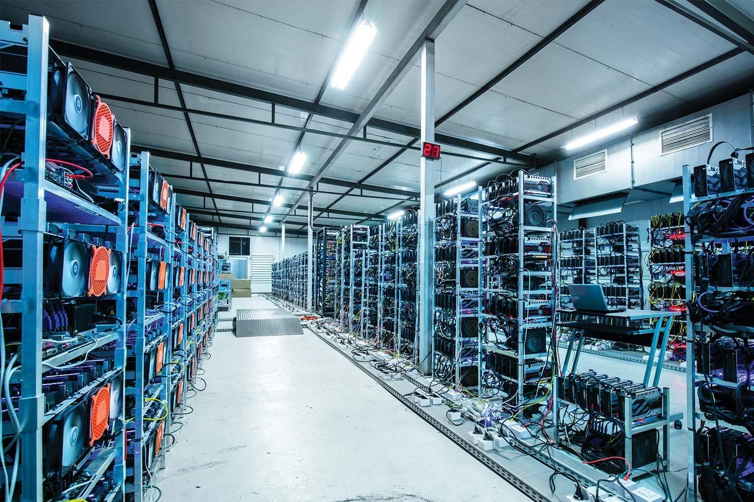 Rows of metal racks holding wiry, complex computer equipment that mine bitcoin sit in a warehouse lit by fluorescent lights.