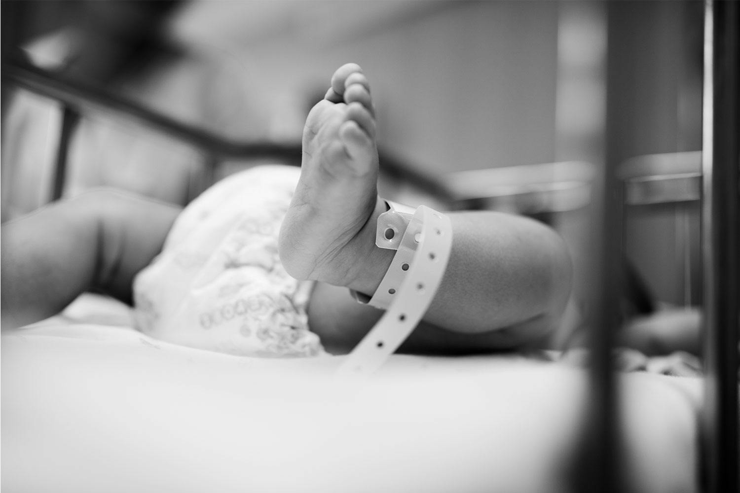 A newborn baby lies in a hospital crib. The baby has a plastic snap-button fastened anklet on their left leg.