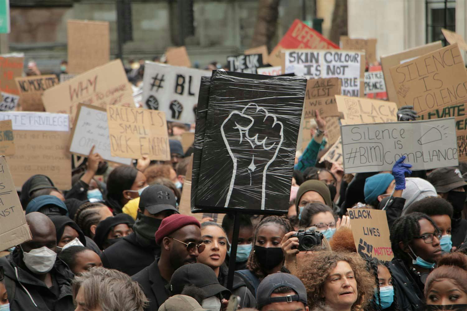 A crowd of BLM protesters hold signs. In the middle, a black cardboard sign shows a fist held upward, drawn in white paint.