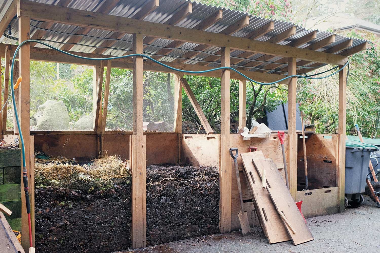 A wooden shed with a corrugated metal roof. The shed's four sections hold piles of dark brown compost about 1 metre high.