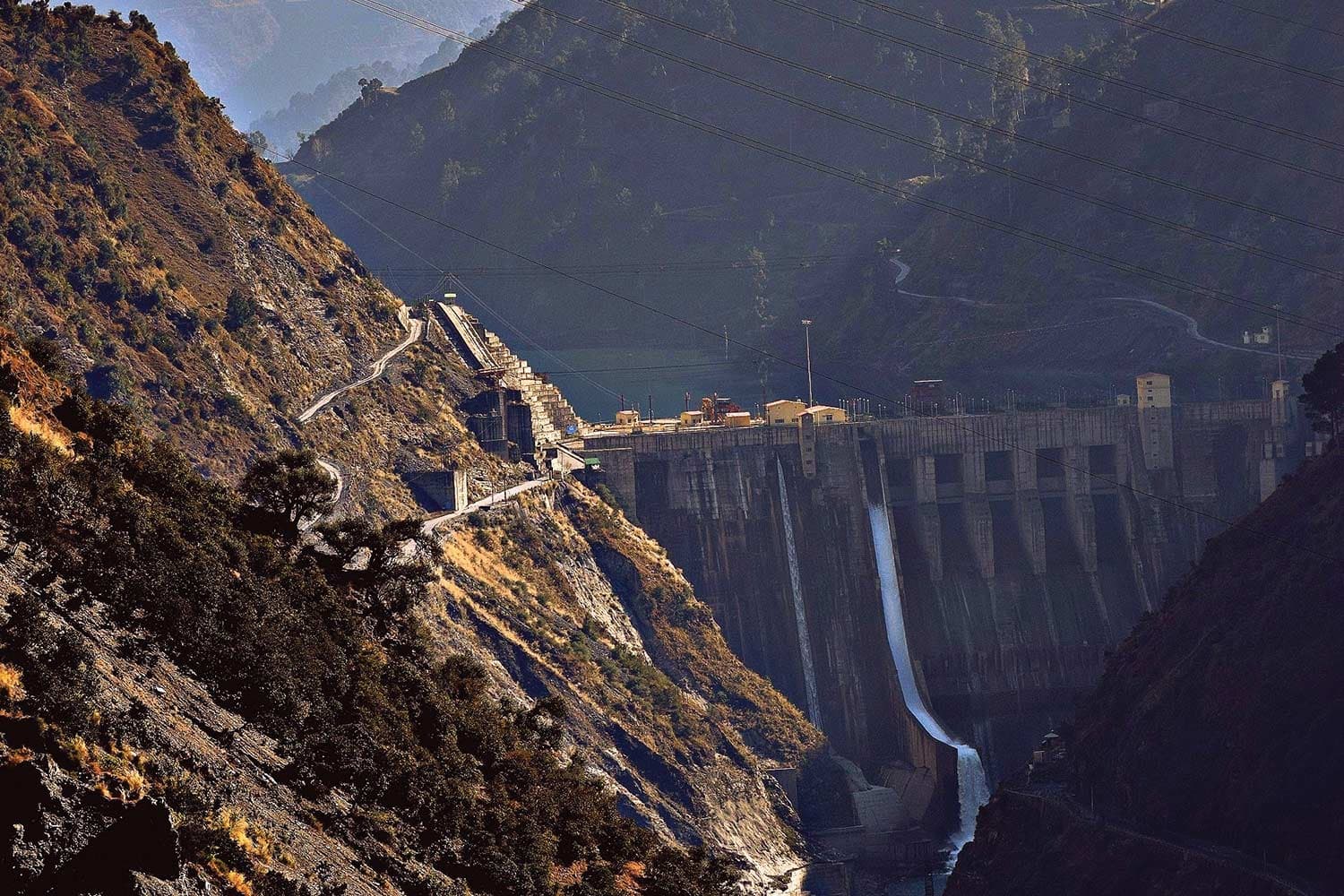 A concrete dam sits in a valley in Kashmir. The sunlit mountain to the left is rocky and dotted with golden grass and trees.