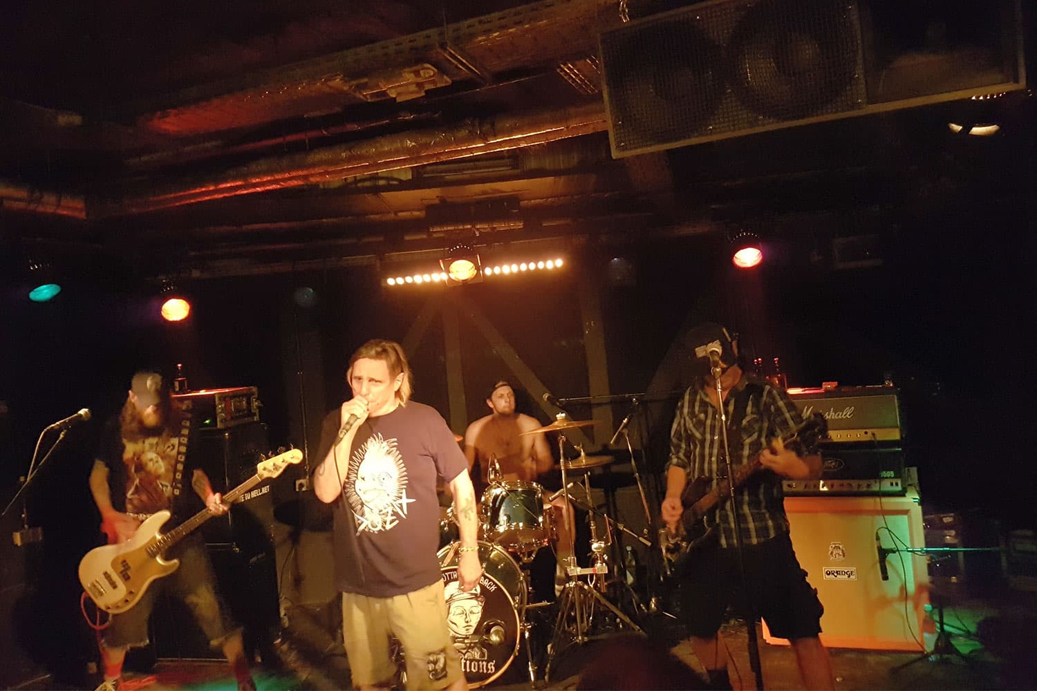 A 4-member punk band wearing dark clothes perform on a stage lit by three orange spotlights. A big speaker hangs from above.