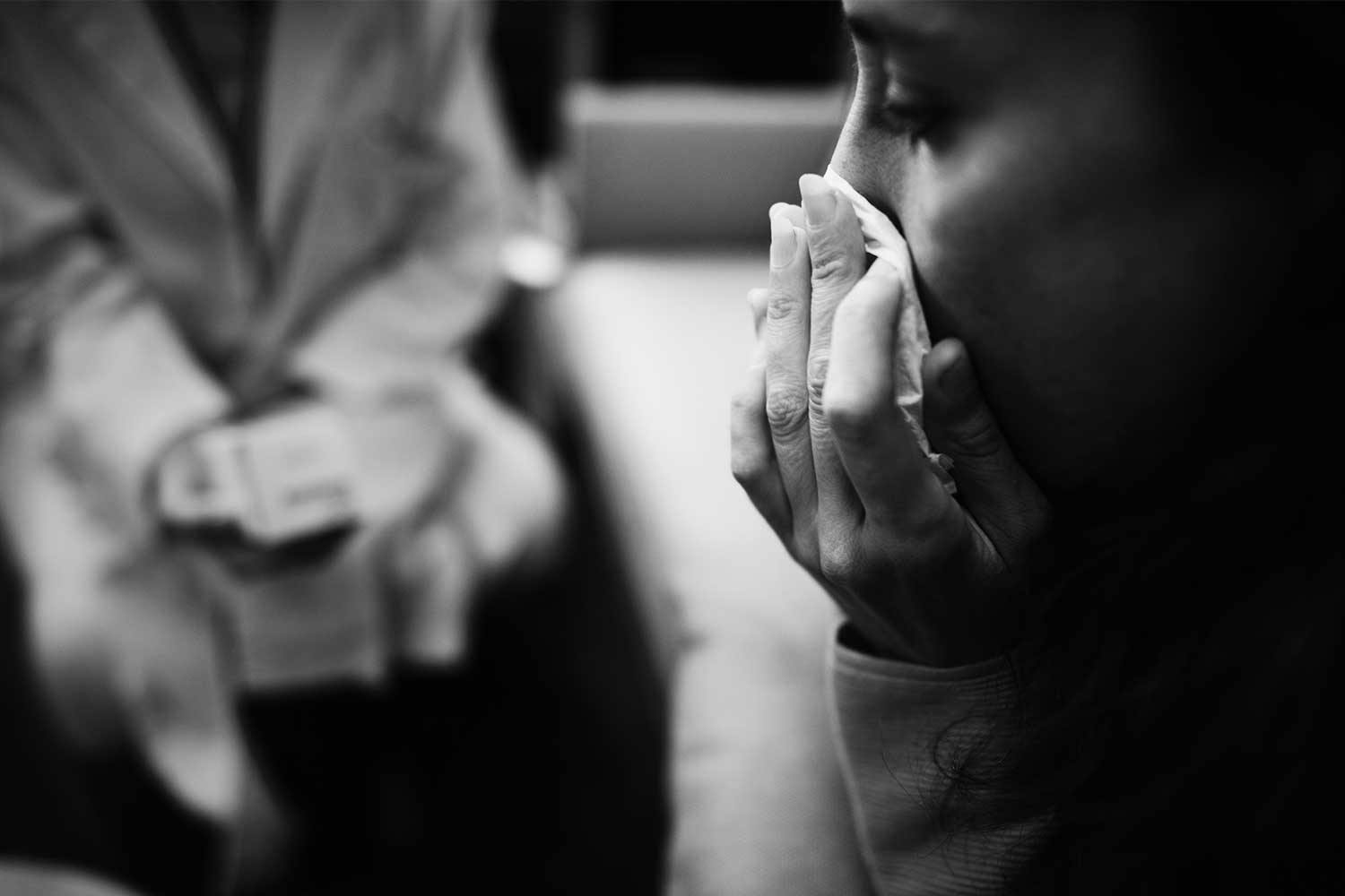 A woman sitting across from a doctor wipes her nose with a tissue.