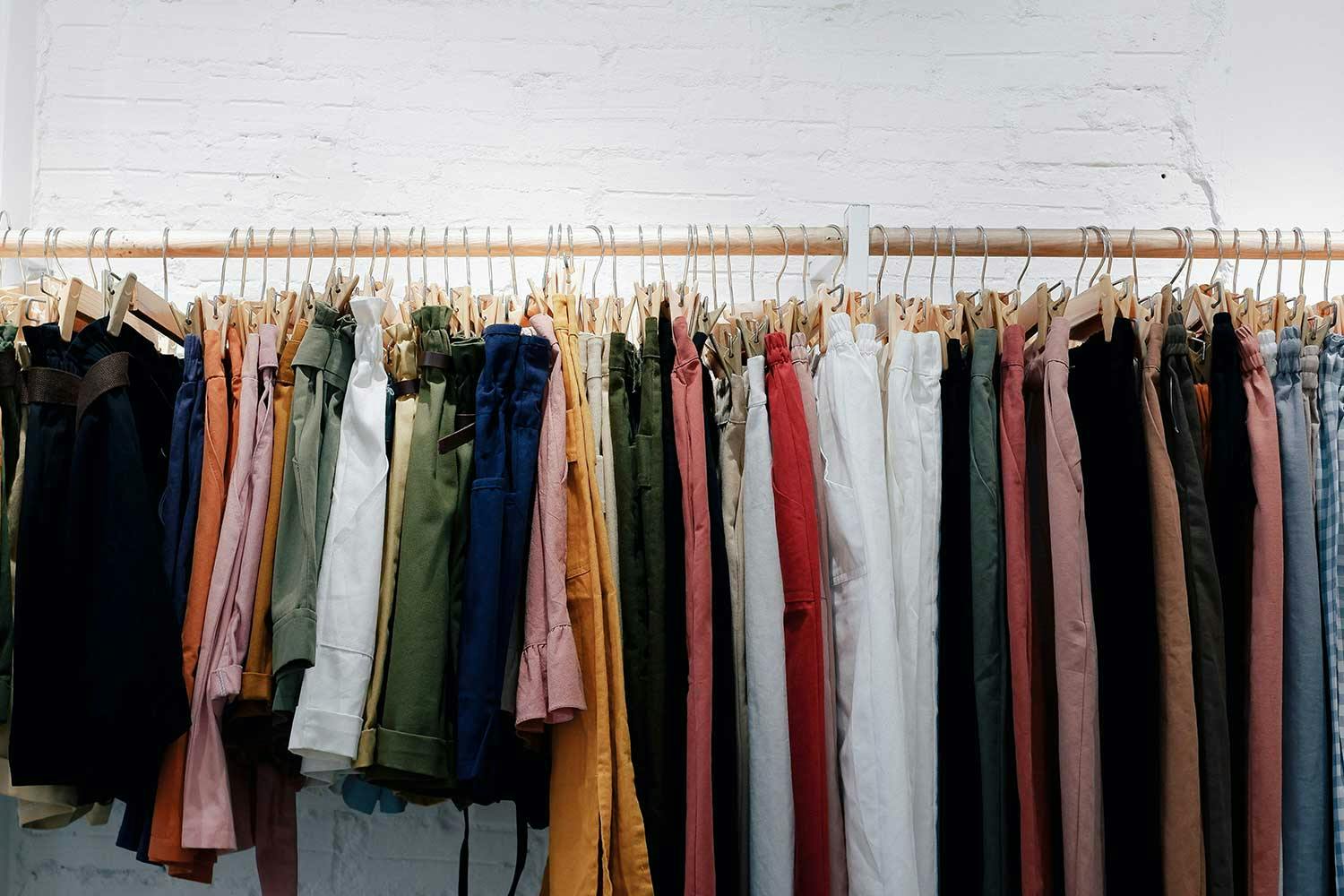 A rack of shorts, skirts, and pants of various styles and colours on pants hangers, against a white brick wall.