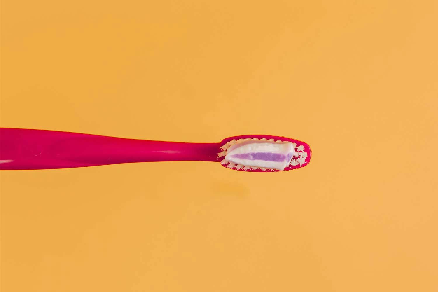 A dab of purple and white toothpaste on the bristles of a pink toothbrush, on a yellow background.
