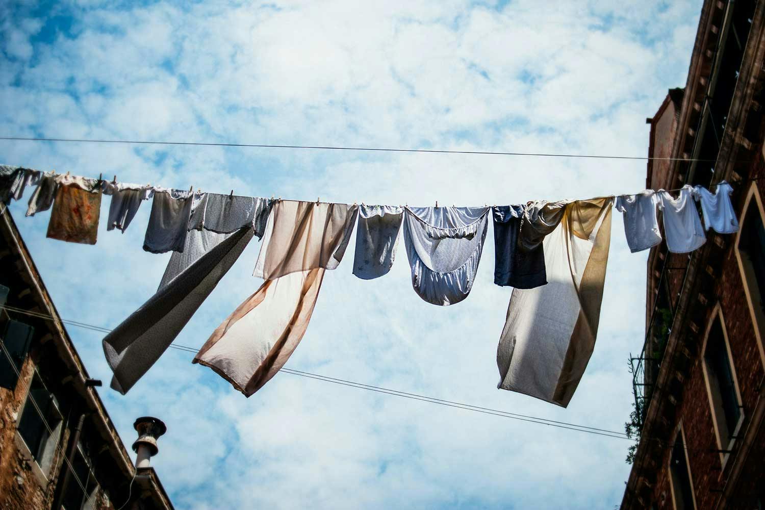 T-shirts and bed linens hang on a line stretched between two old buildings, under a bright blue sky with fluffy clouds.