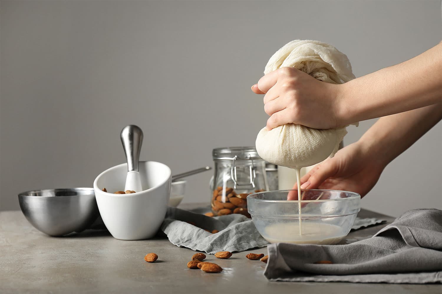 A person strains almond milk through a cloth into a glass bowl next to a jar of almonds, a metal bowl, & a mortar & pestle.