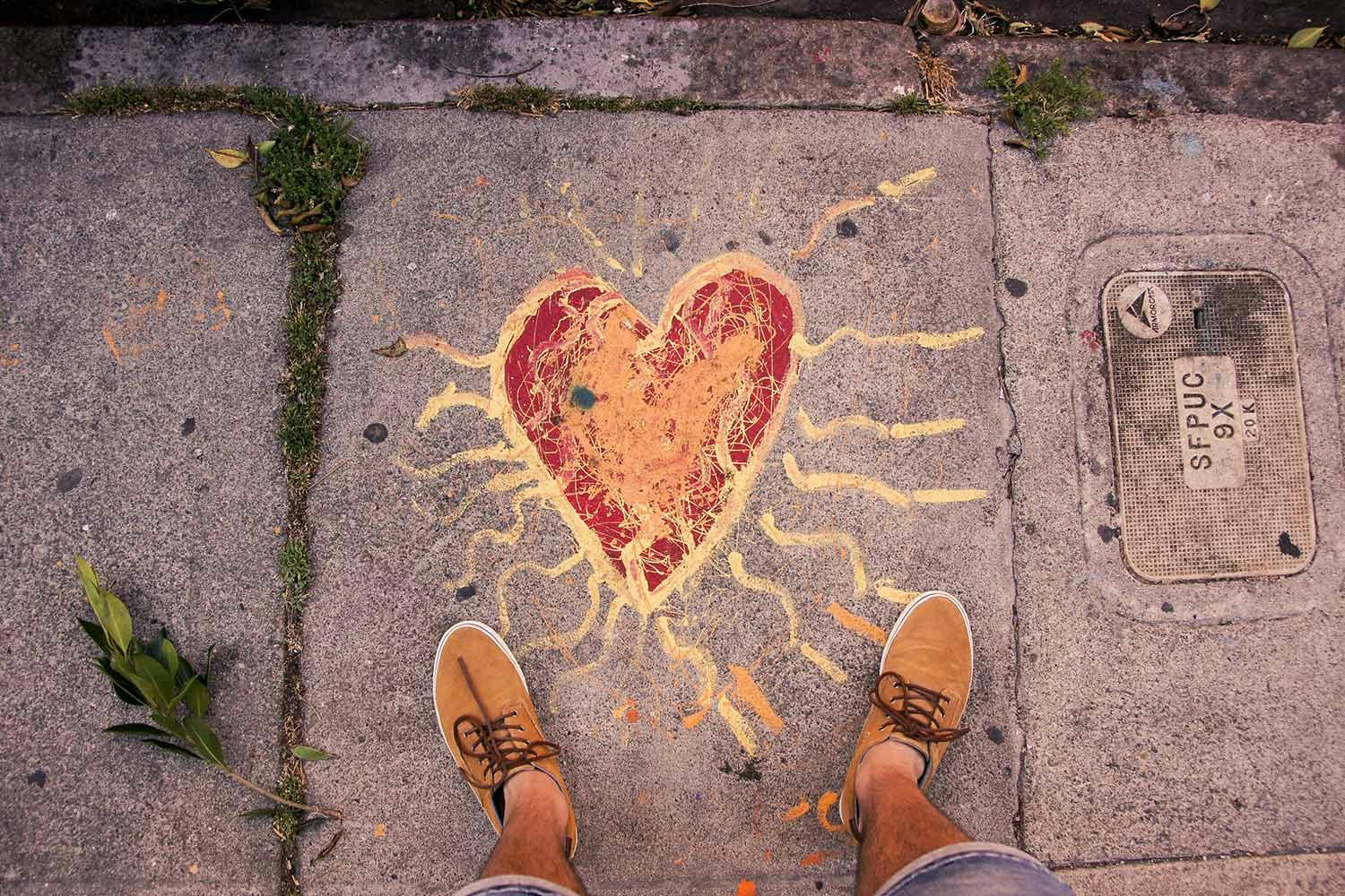 A person wearing brown shoes stands at a red heart with yellow lines radiating from it drawn on a concrete sidewalk.