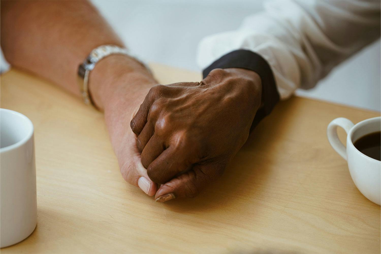 Two people, one light skinned and one dark skinned, holding hands on a wooden table. Next to them are cups of coffee.