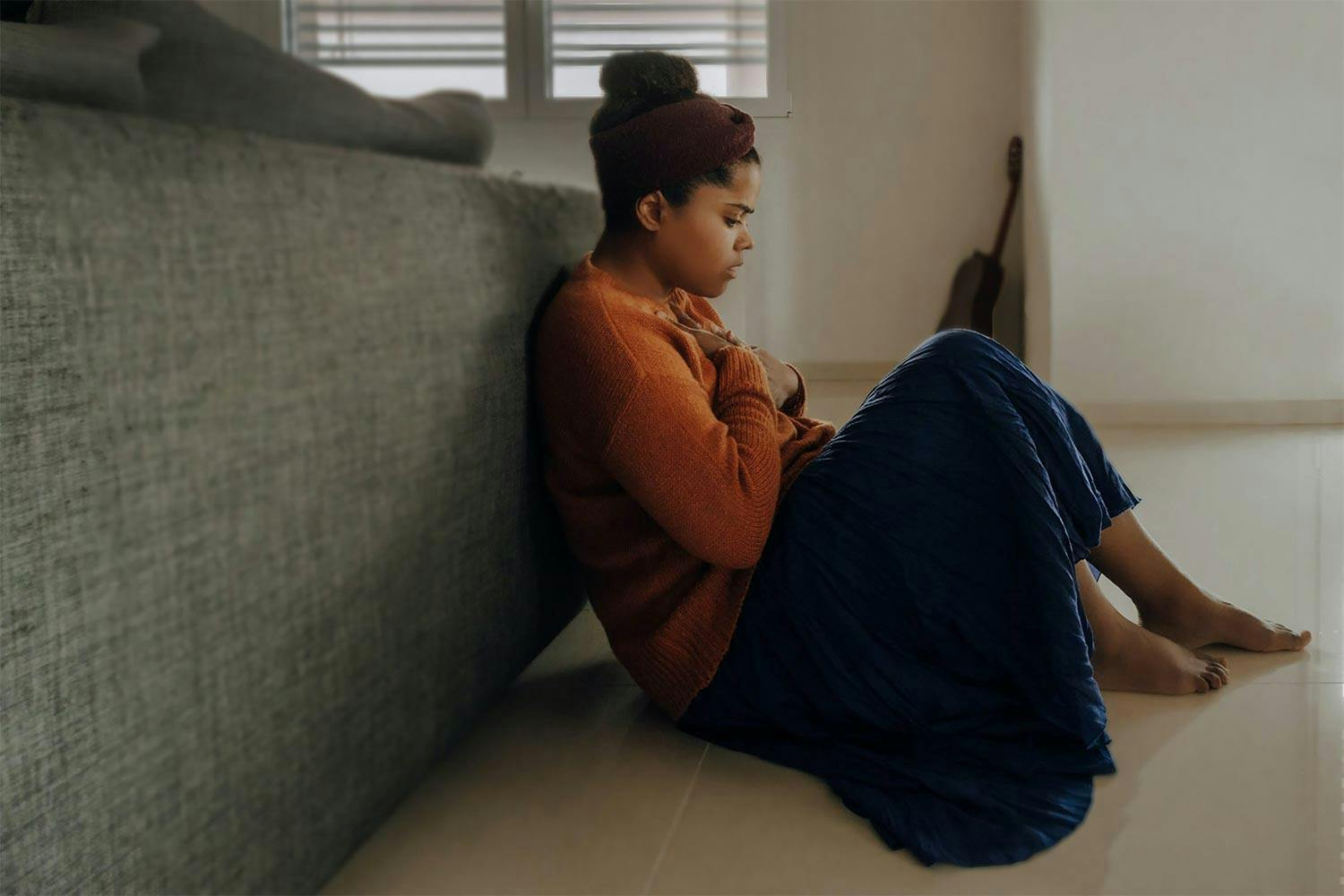A young Black woman sits on the floor leant against the back of a couch holding her chest with both hands looking distressed.