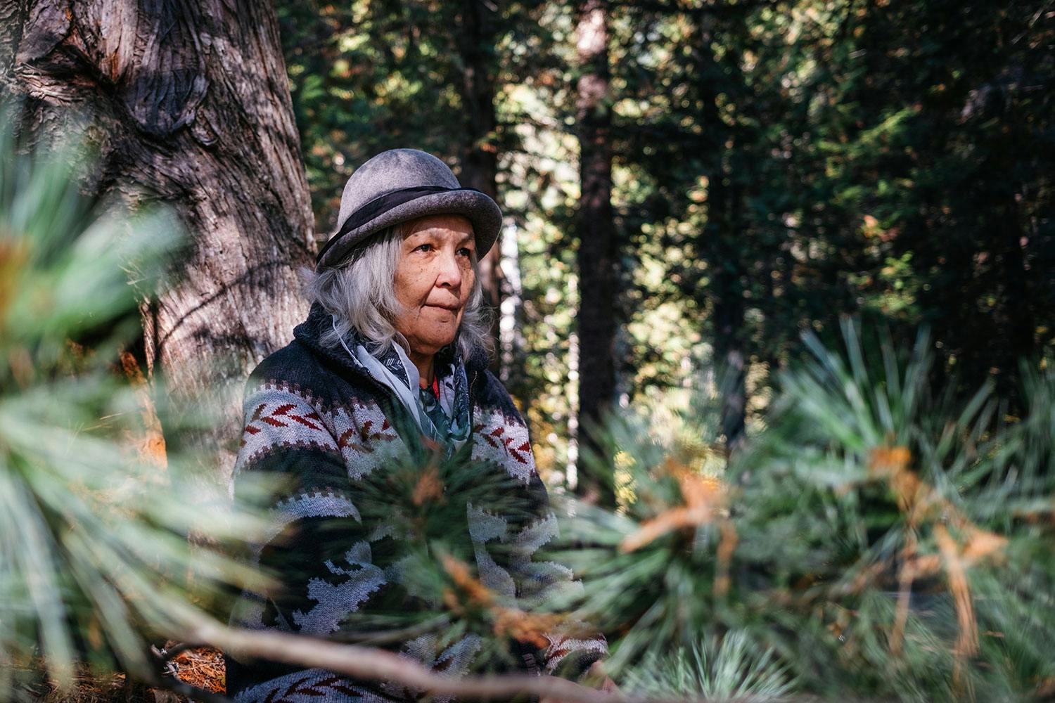 Marilyn James, a Sinixt woman with tan skin & medium grey hair wears a grey hat and grey patterned sweater, sits in a forest.
