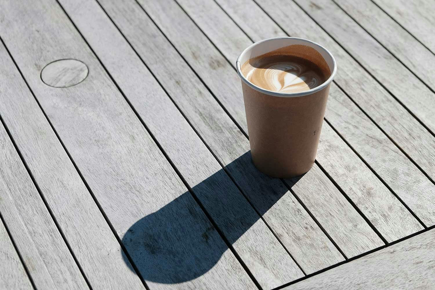 A latte in a disposable coffee cup sitting on a wooden patio table. Sunlight casts a shadow of the cup.