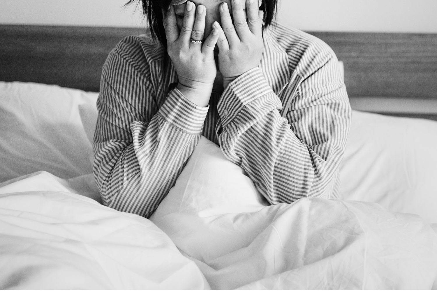 A woman buries her face in her hands. She wears a striped button-down shirt and sits in bed.